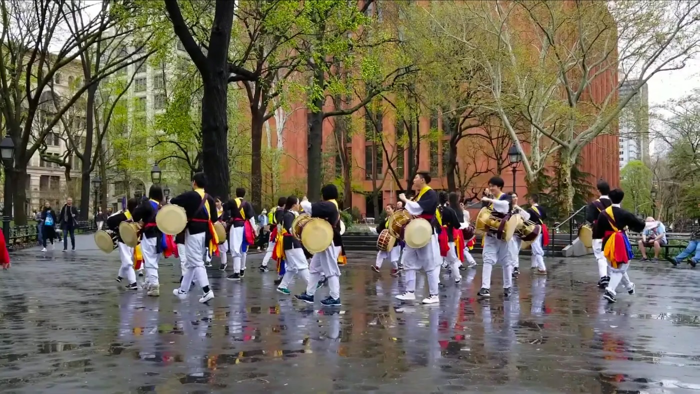 Members drumming and dancing in a pungmul procession on a rain-wet plaza in Washington Square Park